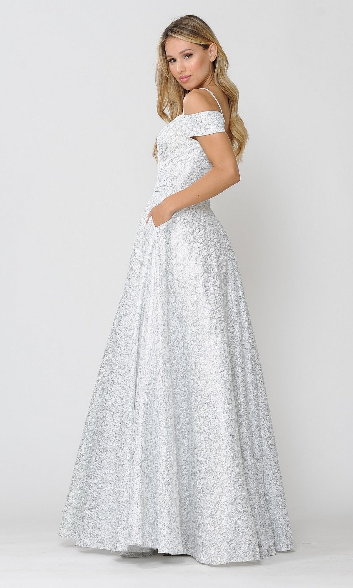 A woman stands in profile against a plain light background, wearing the Cold-Shoulder Silver and White Long Prom Gown 8380 with off-shoulder detailing, a textured finish, and her hand tucked in a pocket.